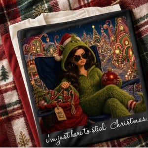 May include: A black t-shirt featuring a graphic of a person in a green fuzzy hooded outfit and sunglasses, holding a red ornament. The scene includes a red sack labeled WHO-VILLE, a blue folding chair, and a festive background with the text I'm just here to steal Christmas.