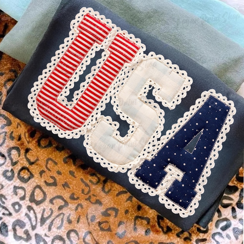 Puede incluir: Un textil azul marino con las letras "USA" aplicadas en telas a rayas rojas y blancas, a cuadros y azul marino con lunares. Cada letra est&aacute; bordeada con un ribete de encaje blanco. El textil est&aacute; sobre una superficie con estampado de leopardo.
