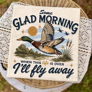 May include: A square, off-white canvas print featuring a vintage-style illustration of a flying duck. The artwork includes the text "Some Glad Morning When This Life Is Over I'll Fly Away" in navy blue script. The duck has brown, blue, and green feathers.