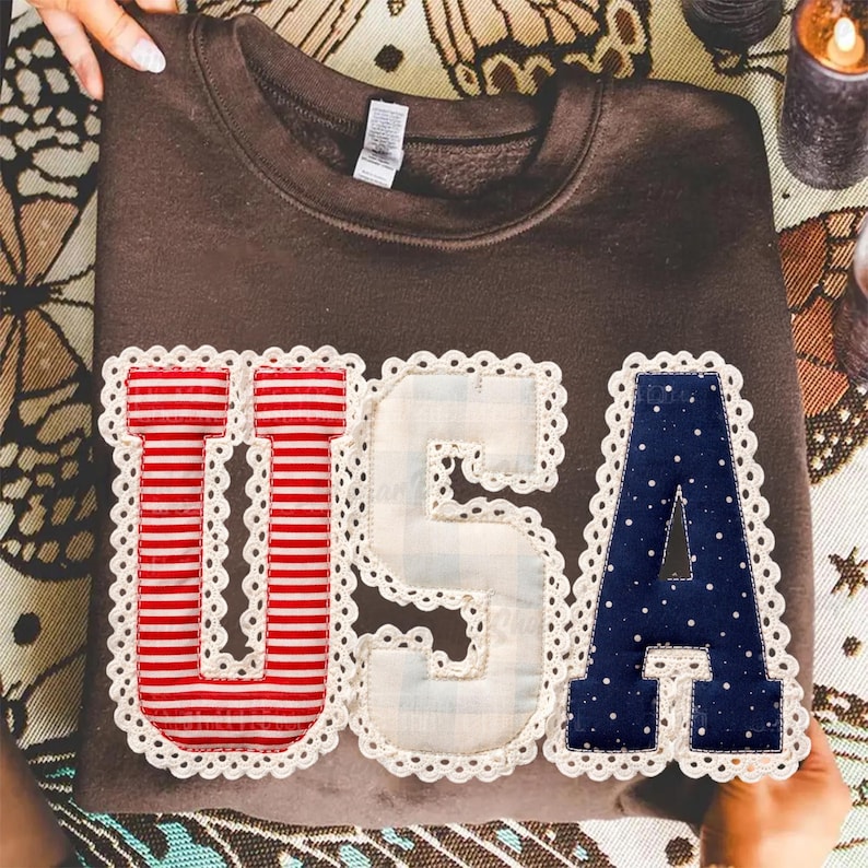Puede incluir: Una sudadera marr&oacute;n con la palabra "USA" aplicada en la parte delantera. Las letras est&aacute;n decoradas con rayas rojas y blancas, un dise&ntilde;o de lunares azules y un patr&oacute;n de cuadros blancos, todo ello ribeteado con encaje blanco.
