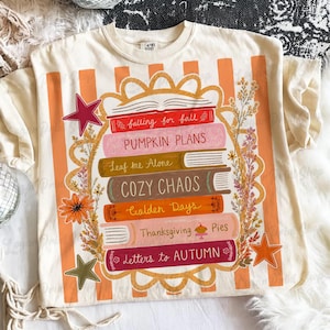 Puede incluir: Una camiseta de color crema con un gr&aacute;fico de una pila de seis libros con los t&iacute;tulos falling for fall, PUMPKIN PLANS, Leaf me Alone, COZY CHAOS, Golden Days, Thanksgiving Pies y Letters to AUTUMN, rodeados de motivos florales y estrellas.
