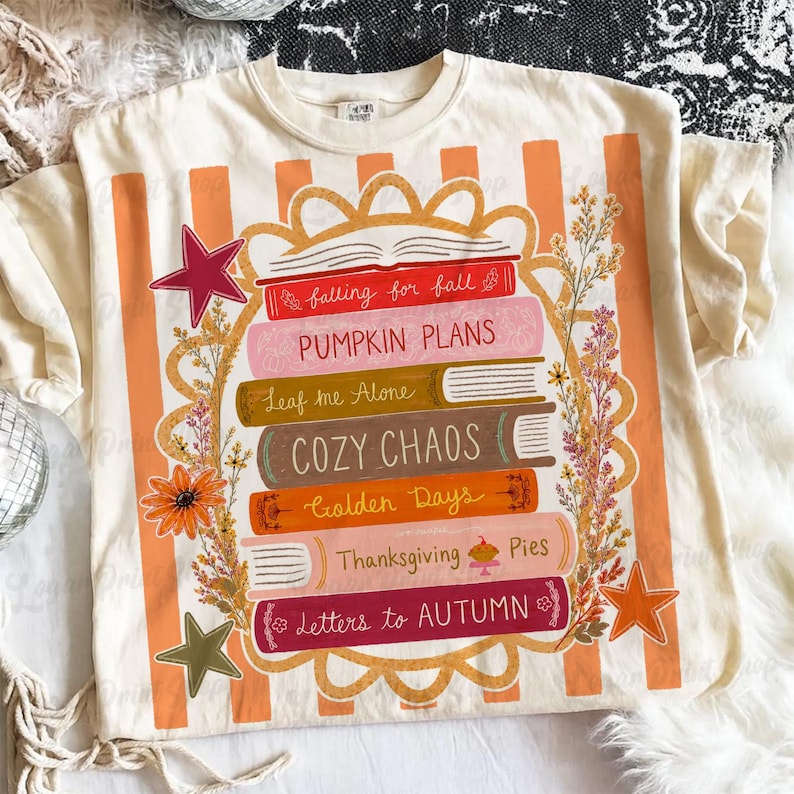 Puede incluir: Una camiseta de color crema con un gr&aacute;fico de una pila de seis libros con los t&iacute;tulos falling for fall, PUMPKIN PLANS, Leaf me Alone, COZY CHAOS, Golden Days, Thanksgiving Pies y Letters to AUTUMN, rodeados de motivos florales y estrellas.