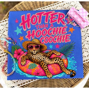 Puede incluir: Camiseta azul con el texto rosa "Hotter Than A Hoochie Coochie". El dise&ntilde;o presenta un leopardo con gafas de sol y sombrero de paja, descansando en un flotador inflable rosa. Estrellas y flores tropicales.