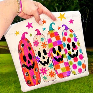 May include: A white t-shirt featuring a graphic print of four tall, colorful pumpkin-shaped ghosts. Each ghost has a unique pattern including stripes with flowers, checkered squares, and polka dots, accented by small colorful stars above them.