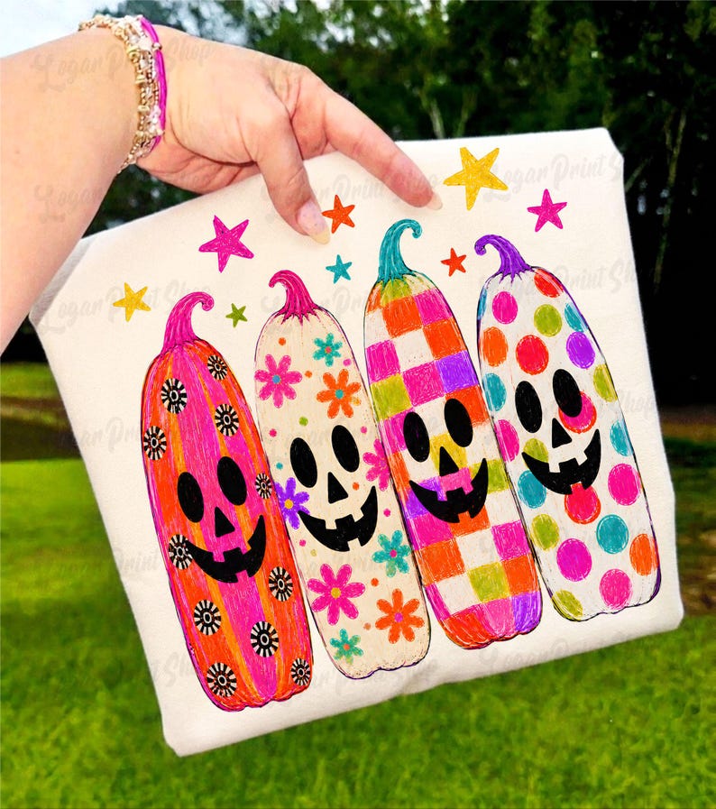May include: A white t-shirt featuring a graphic print of four tall, colorful pumpkin-shaped ghosts. Each ghost has a unique pattern including stripes with flowers, checkered squares, and polka dots, accented by small colorful stars above them.