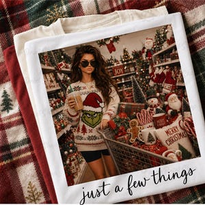 May include: A white t-shirt featuring a graphic print of a person wearing a Grinch-themed sweater and holding a drink in a store filled with Christmas decorations. The text "just a few things" is printed in black cursive script at the bottom.