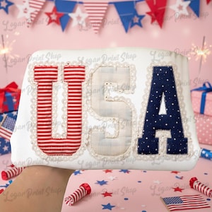 Puede incluir: Un coj&iacute;n blanco con las letras "USA" aplicadas. La "U" es a rayas rojas y blancas, la "S" es a cuadros azul claro y la "A" es azul marino con lunares blancos. Las letras est&aacute;n bordeadas con encaje blanco.