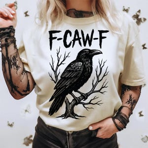 May include: Cream-colored t-shirt featuring a black raven perched on a tree branch. The text "F-CAW-F" is above the raven. The shirt has short sleeves and is worn with black lace arm sleeves.