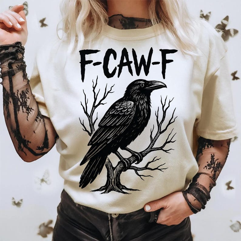 May include: Cream-colored t-shirt featuring a black raven perched on a tree branch. The text "F-CAW-F" is above the raven. The shirt has short sleeves and is worn with black lace arm sleeves.