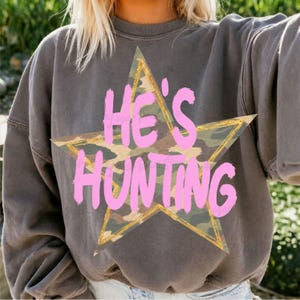 May include: A charcoal gray crewneck sweatshirt featuring a large camouflage-patterned star graphic with a gold outline. The text HE'S HUNTING is printed across the center of the star in a bold, bright pink, hand-lettered style font.