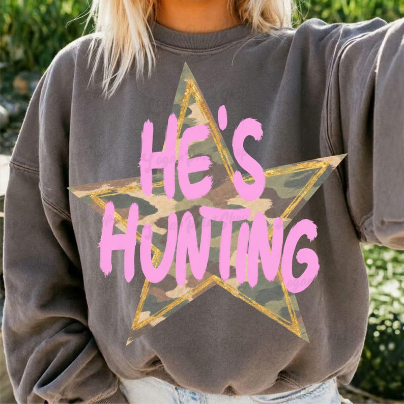 May include: A charcoal gray crewneck sweatshirt featuring a large camouflage-patterned star graphic with a gold outline. The text HE'S HUNTING is printed across the center of the star in a bold, bright pink, hand-lettered style font.