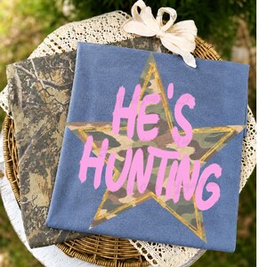 May include: A blue t-shirt featuring a large star with a camouflage pattern and a gold outline. The text HE'S HUNTING is printed in a bright pink, brush-stroke font across the center of the star. A second camouflage-patterned fabric is visible.