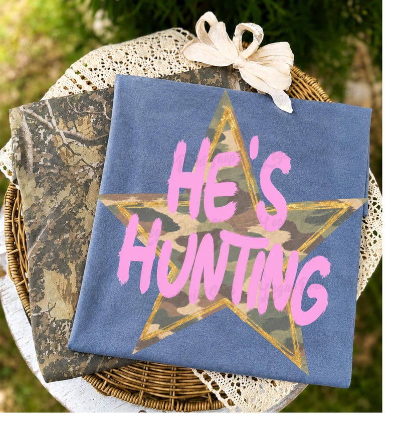 May include: A blue t-shirt featuring a large star with a camouflage pattern and a gold outline. The text HE'S HUNTING is printed in a bright pink, brush-stroke font across the center of the star. A second camouflage-patterned fabric is visible.