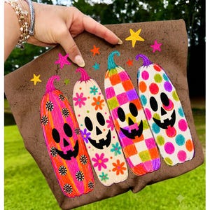 May include: A brown fabric pouch featuring a colorful graphic of four tall, slender pumpkins with jack-o'-lantern faces. The pumpkins are decorated with bright floral, checkered, and polka dot patterns in pink, orange, green, and blue, with small stars.