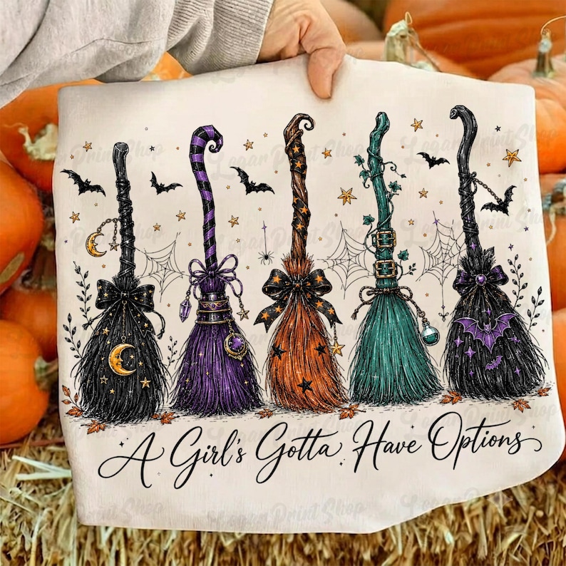 May include: A white t-shirt featuring a graphic of five stylized witch brooms in black, purple, orange, and teal, decorated with stars, moons, bats, and spiderwebs, with the text A Girl's Gotta Have Options printed in black script at the bottom.