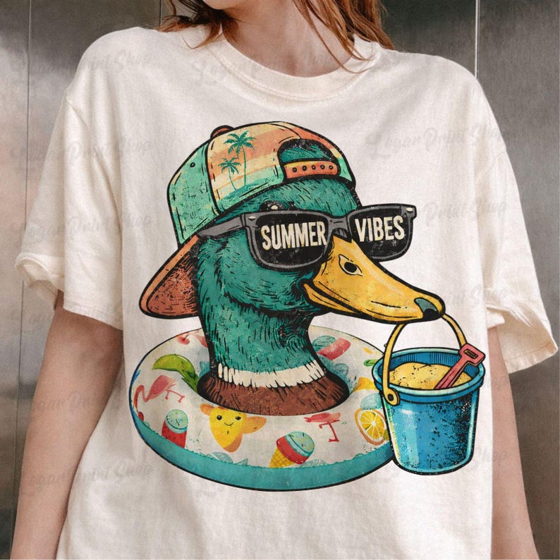 May include: Cream-colored t-shirt featuring a cartoon duck wearing sunglasses that read "Summer Vibes." The duck is wearing a hat and a floatie, holding a bucket with sand and a shovel. The design has a beach theme.
