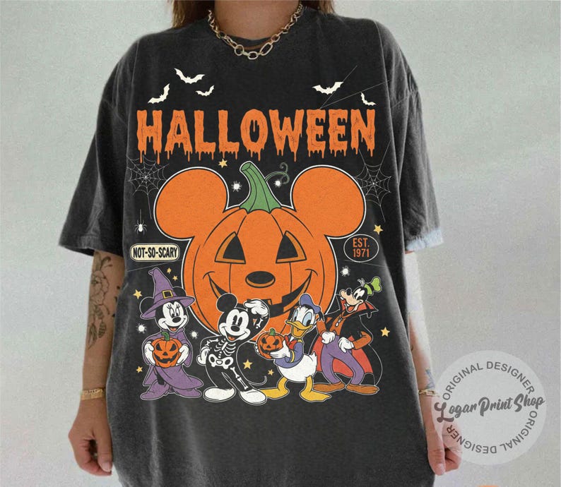 May include: Dark gray Halloween-themed t-shirt featuring a large orange pumpkin with Mickey Mouse ears and the word "HALLOWEEN" in orange. The shirt also features Mickey Mouse, Donald Duck, Goofy, and Minnie Mouse in Halloween costumes.