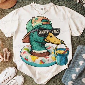 May include: White t-shirt featuring a cartoon duck wearing sunglasses that read "Summer Vibes" and a hat. The duck is in a pool floatie and holding a bucket. The shirt has a beach theme.