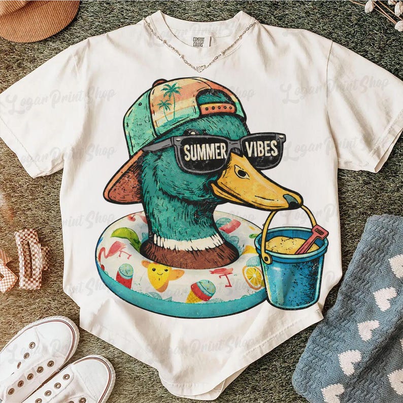 May include: White t-shirt featuring a cartoon duck wearing sunglasses that read "Summer Vibes" and a hat. The duck is in a pool floatie and holding a bucket. The shirt has a beach theme.