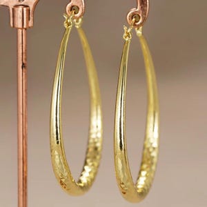 May include: A pair of gold-colored hoop earrings. The earrings have a hammered texture and are displayed on a copper-colored earring stand. The hoops are oval-shaped and have a shiny finish. The background is a soft, neutral color.
