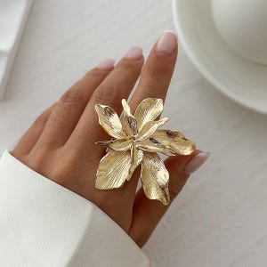 May include: A gold-toned statement ring featuring a large, detailed flower design. The ring is made of metal and has a textured finish.