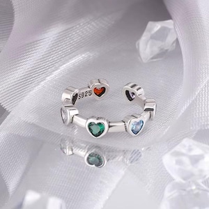 May include: Silver ring with a row of five heart-shaped stones. The stones are red, green, blue, purple, and clear. The ring is marked "S925".