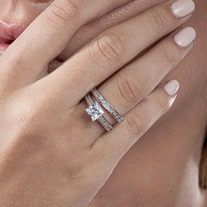 May include: A silver ring set with a square, clear gemstone as the focal point. The band is embellished with smaller, clear gemstones. The ring is displayed on a finger, showcasing the design and sparkle of the jewellery.
