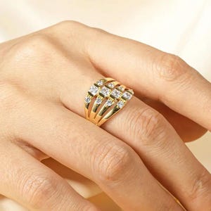 May include: A gold-colored ring with multiple bands, each adorned with small, clear gemstones. The ring is worn on a finger, showcasing its intricate design and sparkling details. The ring's design is elegant and eye-catching.