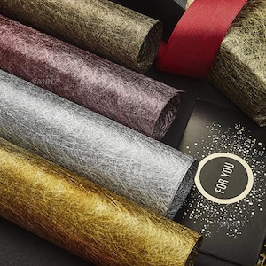 May include: Close-up of rolled decorative paper in gold, silver, burgundy, and brown. A gift box with a red ribbon and a black card with the text "FOR YOU" are also visible. The paper has a textured, fibrous appearance.