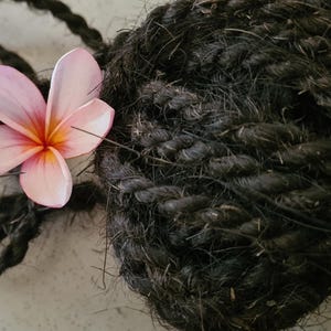 Op de afbeelding: Close-up van een donkerbruine, getextureerde touwbal met een roze en gele plumeria bloem. De bloem bevindt zich aan de linkerkant van de afbeelding, de touwbal vult de rechterkant. Het touw lijkt strak gewikkeld.