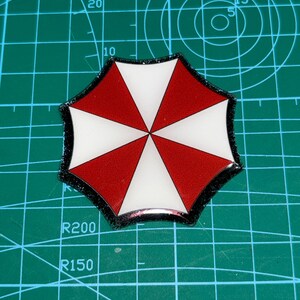 May include: A red, white, and black umbrella-shaped emblem. The emblem features a segmented design with alternating red and white triangular sections. The outer edge is outlined in black. The emblem is set against a green grid background.
