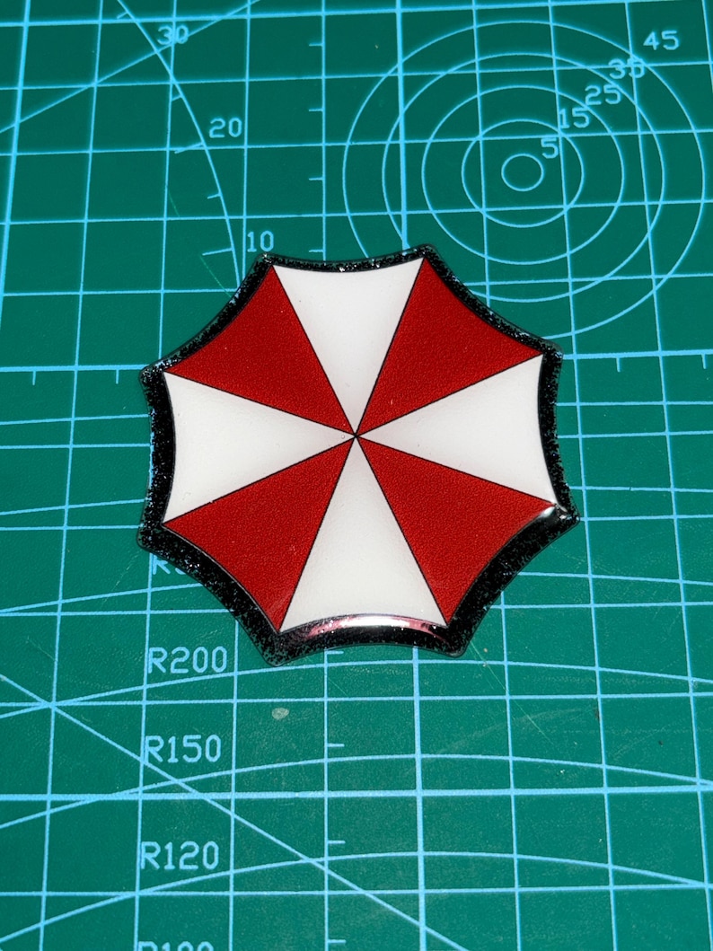 May include: A red, white, and black umbrella-shaped emblem. The emblem features a segmented design with alternating red and white triangular sections. The outer edge is outlined in black. The emblem is set against a green grid background.