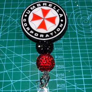 May include: A black and white badge reel with the text "UMBRELLA CORPORATION" around a red and white umbrella logo. The reel has black and red beaded accents and a clear plastic clip.