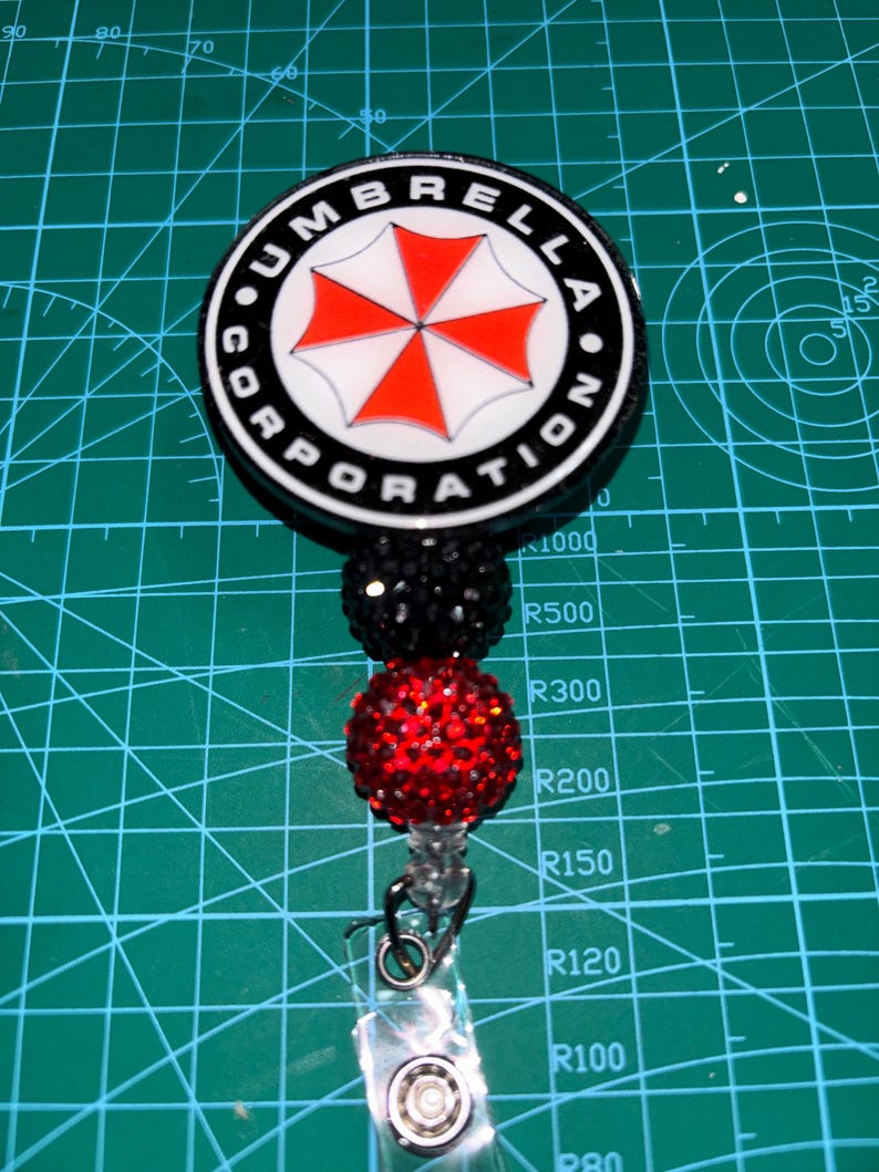 May include: A black and white badge reel with the text "UMBRELLA CORPORATION" around a red and white umbrella logo. The reel has black and red beaded accents and a clear plastic clip.