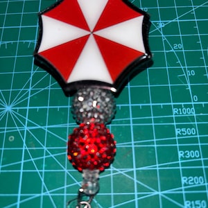 May include: A badge reel featuring a red, white, and black umbrella design. The reel has a black border, with a red and white segmented pattern. The reel is attached to a beaded accent and a clear plastic clip.