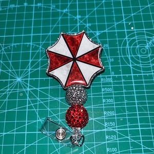 May include: A badge reel featuring a red, white, and black umbrella design. The badge reel has a silver glittery top, a red glittery ball, and a clear plastic clip. The design is on a green cutting mat.