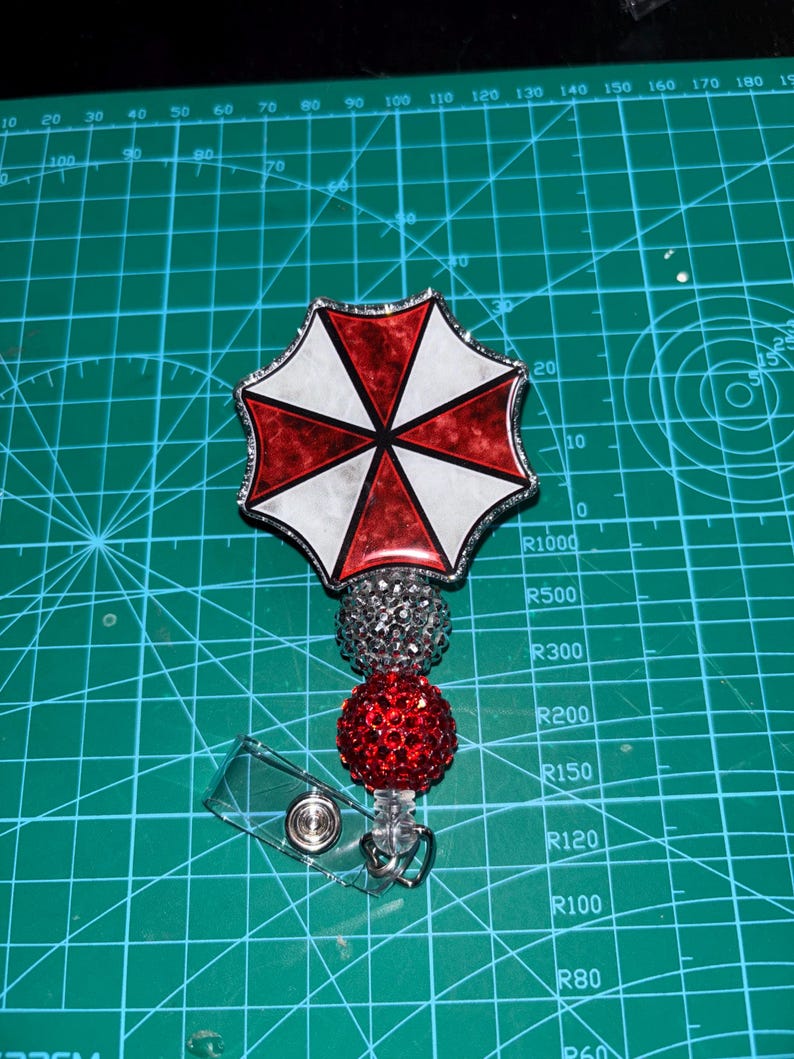 May include: A badge reel featuring a red, white, and black umbrella design. The badge reel has a silver glittery top, a red glittery ball, and a clear plastic clip. The design is on a green cutting mat.