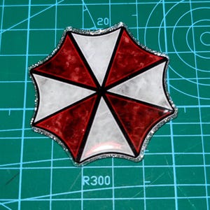May include: A resin-based, octagonal emblem featuring a red, white, and black color scheme. The design is a segmented umbrella shape with a silver glitter border. The emblem is set against a green cutting mat.