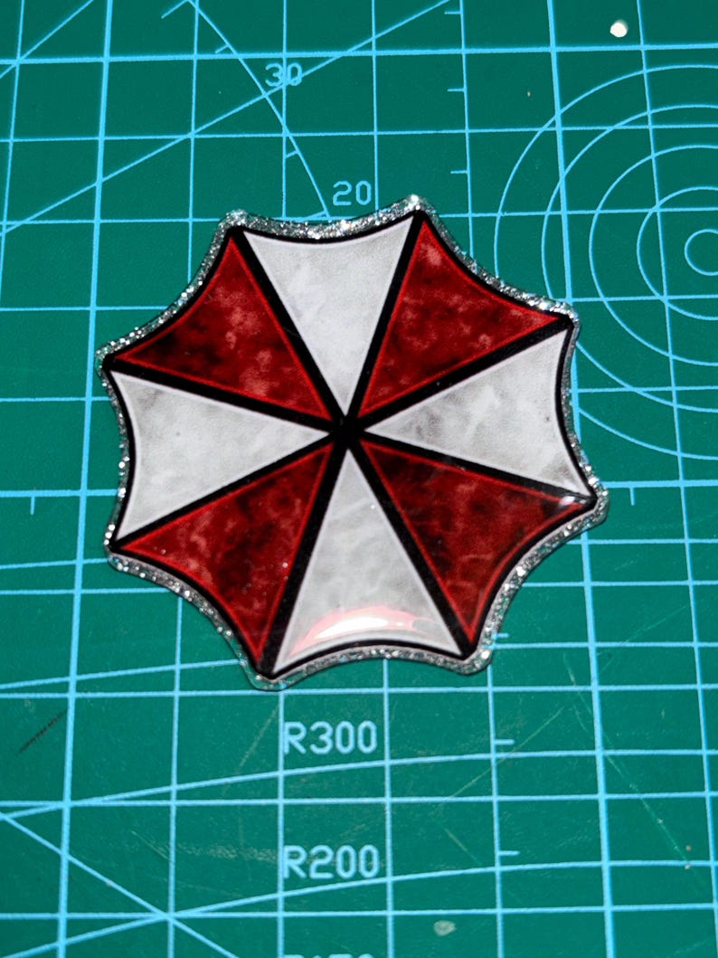May include: A resin-based, octagonal emblem featuring a red, white, and black color scheme. The design is a segmented umbrella shape with a silver glitter border. The emblem is set against a green cutting mat.
