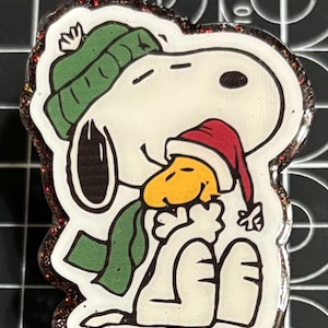 May include: A Snoopy and Woodstock-themed badge reel. Snoopy is wearing a green knit cap and scarf, and Woodstock is wearing a red Santa hat. The badge reel has a glittery, brown border.