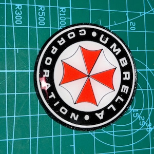 May include: A round sticker with the text "UMBRELLA CORPORATION" in white lettering on a black border. The center features a red and white umbrella logo. The sticker is on a green cutting mat.