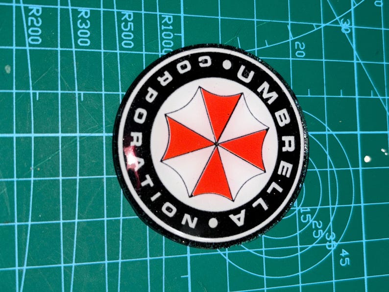 May include: A round sticker with the text "UMBRELLA CORPORATION" in white lettering on a black border. The center features a red and white umbrella logo. The sticker is on a green cutting mat.