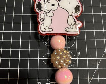May include: A Snoopy-themed retractable badge reel featuring Snoopy and a female dog in a kissing pose, with a pink background and red glitter trim. The reel includes pink and gold beads and a clear plastic clip.