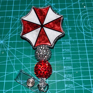 May include: A retractable badge reel featuring a red, white, and black umbrella logo. The badge reel has a silver glitter ball and a red glitter ball. The reel is attached to a clear plastic clip.