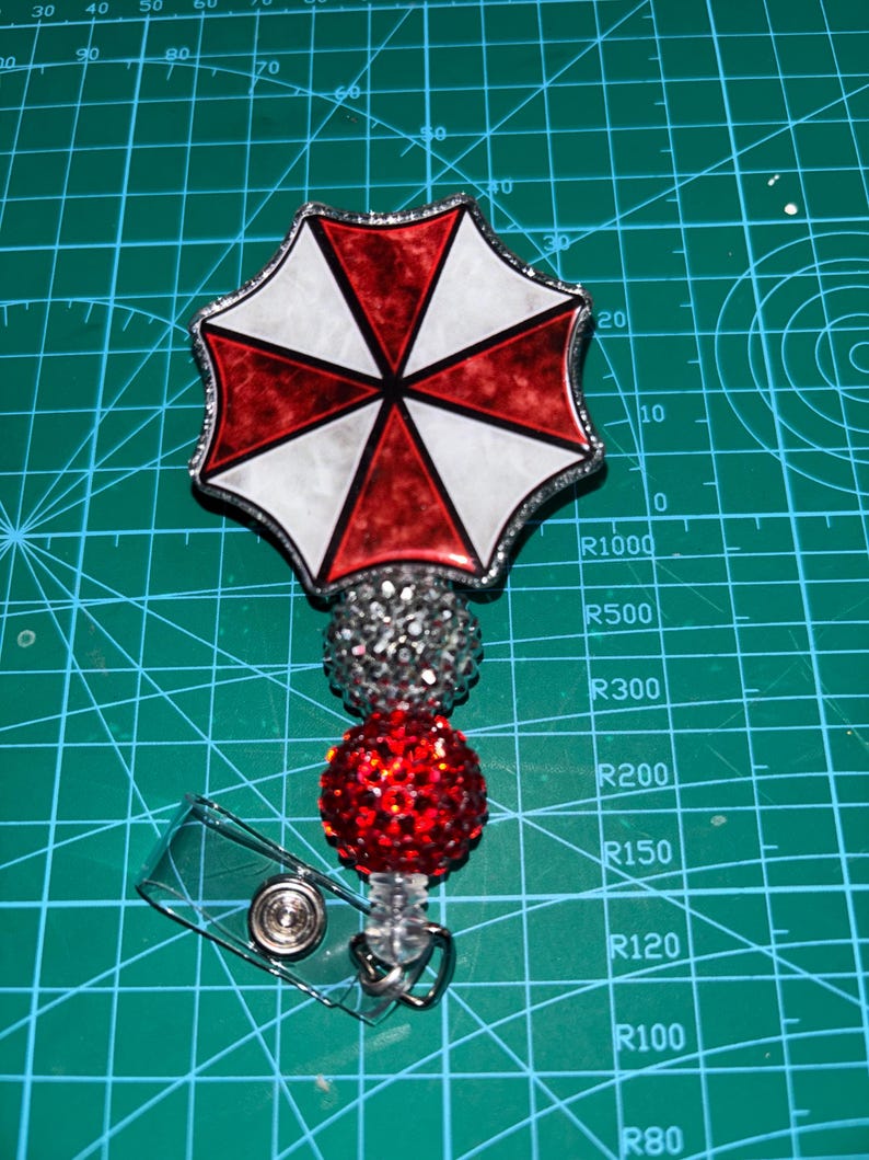 May include: A retractable badge reel featuring a red, white, and black umbrella logo. The badge reel has a silver glitter ball and a red glitter ball. The reel is attached to a clear plastic clip.