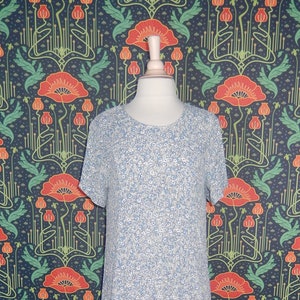 May include: A light blue floral print dress with short sleeves and a tiered skirt is displayed on a mannequin. The dress features a round neckline and a delicate pattern of small white and blue flowers. The mannequin is set against a backdrop of a floral wallpaper.