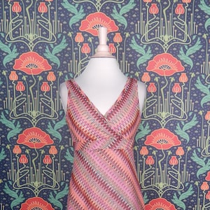 May include: A sleeveless, V-neck dress with a pink, orange, and brown chevron pattern. The dress is displayed on a pink dress form against a dark blue floral wallpaper background.