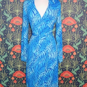 May include: A blue dress with a leaf pattern is displayed on a mannequin. The dress has long sleeves, a V-neck, and a matching belt. The background features a floral wallpaper with red and orange flowers.