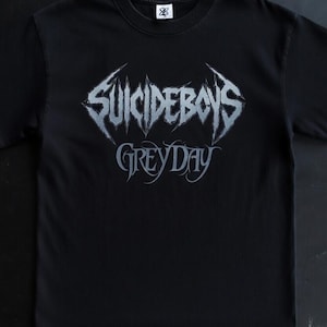 May include: Black t-shirt with the band name "SUICIDEBOYS" in a metallic font above "GREYDAY" in a gothic script. The dark fabric tee has a crew neck. A small logo is visible on the collar.