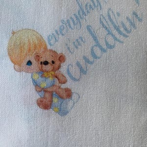 May include: A light blue fabric with a Precious Moments illustration. The image features a blonde-haired child holding a teddy bear, with the text "everyday I'm cuddlin'" in light blue script.